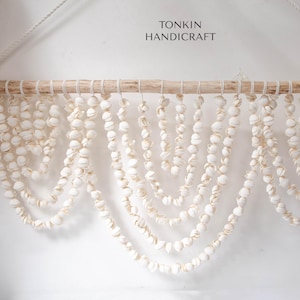 Peut inclure: Décoration murale composée de coquillages blancs suspendus à une baguette en bois naturel. Les coquillages sont enfilés, créant un effet de cascade. Le texte "TONKIN HANDICRAFT" est visible en haut.