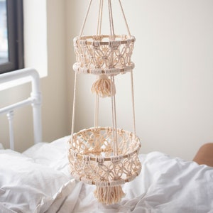 May include: A two-tiered macrame plant hanger with wooden rings and cream-colored cotton cord. The hanger is suspended from a white ceiling and has a tassel at the bottom of each tier.