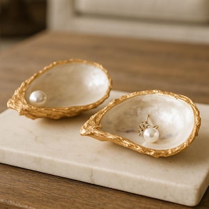 May include: Two decorative oyster shell-shaped dishes with gold edges and a pearlescent interior. One holds a pearl, the other a pearl earring. The text "TONKIN HANDICRAFT" is visible. The dishes are on a white marble surface.