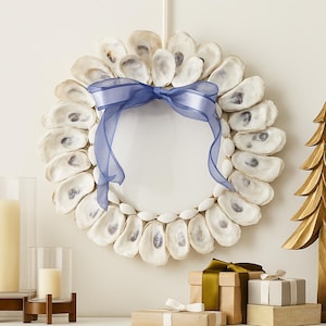 Puede incluir: Una corona redonda hecha de conchas de ostra blancas, adornada con una cinta azul. La corona está rodeada de pequeñas conchas marinas. Un árbol de Navidad de madera dorada y regalos envueltos están sobre una superficie blanca.