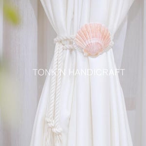 以下が含まれることがあります： 大きな薄いピンク色の貝殻が付いた白いカーテンのタッセル。タッセルは太い白いロープでできており、「TONKN HANDICRAFT」の文字が見えます。カーテンは白です。