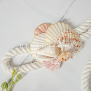 Może przedstawiać: Dekoracyjne ułożenie muszli, w tym muszle wachlarzowate i spiralne, z białą liną i gałązką zielonych liści. Widoczny jest tekst "TONKIN HANDICRAFT". Muszle są w odcieniach kremu, różu i brązu.