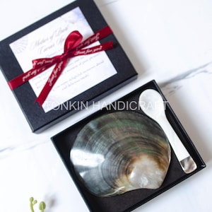 May include: A black gift box tied with a red ribbon, containing a large, iridescent shell and a white spoon. The shell has a pearlescent sheen. The box is on a white marble surface. Text on the box reads "Mother of Pearl".