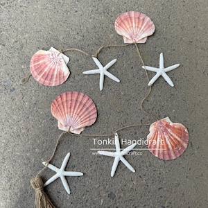 Puede incluir: Guirnalda decorativa de conchas marinas y estrellas de mar. La guirnalda presenta conchas rosas y blancas y estrellas de mar blancas, unidas con hilo. Una borla marrón cuelga del extremo. Ideal para decoración costera.