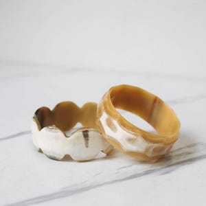 May include: Two horn bracelets, one with a wavy edge and the other with a geometric pattern. Both bracelets are a light brown colour with white and brown markings.