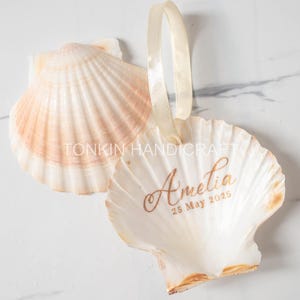 Puede incluir: Dos conchas de vieira, una con el texto "TONKIN HANDICRAFT" y el nombre "Amelia" con la fecha "25 de mayo de 2025" impresos en dorado. Las conchas son de color crema y melocotón, con una cinta crema.