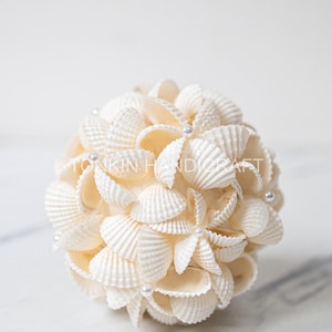 May include: A spherical decorative object made of cream-colored seashells. The shells are arranged in a layered pattern, with small silver beads interspersed throughout. The object is likely a decorative item for a coastal or beach-themed home.