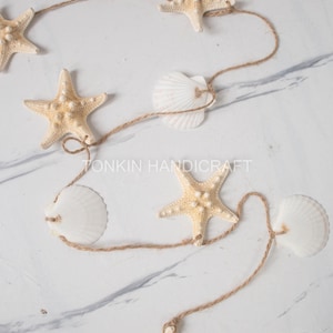 May include: A decorative garland featuring natural starfish and white seashells strung on brown twine. The starfish are beige, and the seashells are white. The garland is arranged on a white marble surface.