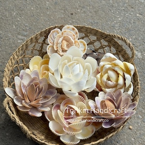 Puede incluir: Una cesta de mimbre llena de flores hechas a mano con conchas marinas en varios colores, incluyendo crema, amarillo y lavanda. Las flores están dispuestas en un patrón circular, mostrando sus intrincados detalles. La cesta está hecha de fibras naturales.