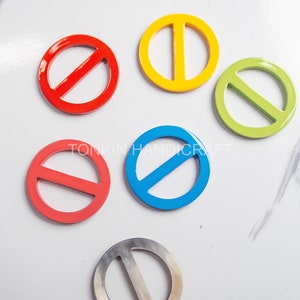 Puede incluir: Seis hebillas de cinturón de metal de colores diferentes: rojo, amarillo, verde, azul, coral y plateado. Cada hebilla es un círculo con una línea horizontal en el centro.