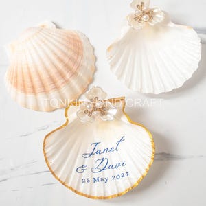 Pode incluir: Três conchas decorativas. Uma concha tem uma borda dourada e o texto azul "Janet & Davi 25 de maio de 2023". Duas conchas têm um enfeite floral com pérolas. As conchas são brancas e pêssego.