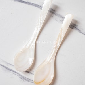 May include: Two white mother of pearl spoons on a white marble surface. The spoons are long and narrow with a rounded bowl.