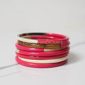 Op de afbeelding: Een stapel van zes kleurrijke armbanden, met een roze, witte en bruine kleurstelling. De armbanden zijn gemaakt van hoorn en zijn op elkaar gestapeld.