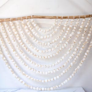 Puede incluir: Adorno de pared con conchas blancas suspendidas de una rama de madera natural. Las conchas están ensartadas, creando un efecto de cascada. Ideal para la decoración del hogar, con un tema costero.