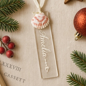 May include: A clear acrylic bookmark with the name "Amelia" in white script, topped with a seashell and tied with a cream ribbon. The bookmark is surrounded by festive decorations, including a wooden star and a red ornament.