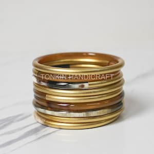 May include: A stack of gold and brown bangles with a white and black pattern. The bangles are stacked on a white marble surface. The text "TONKIN HANDICRAFT" is visible on the bangles.