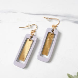 May include: A pair of dangle earrings with a light purple rectangular frame and a gold rectangle inside. The earrings have gold hooks.