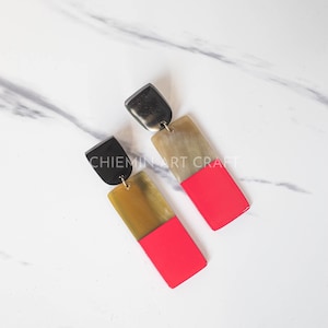 May include: A pair of long, rectangular earrings with a black, brown, and pink color scheme. The earrings are made of horn and have a gold-colored metal connector.