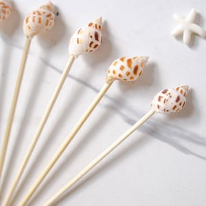 Puede incluir: Brochetas decorativas de conchas marinas con conchas naturales y palitos de madera. Las conchas tienen un patrón moteado blanco y marrón. La imagen también incluye decoraciones de estrellas de mar blancas y el texto "TONKIN HANDICRAFT".