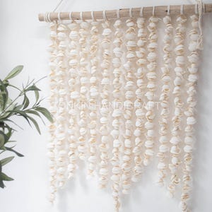 Könnte beinhalten: Wandbehang aus cremefarbenen Muscheln. Viele kleine Muscheln sind an einer natürlichen Holzdübel aufgereiht und aufgehängt. Das Design ist ein Küsten- oder Strandthema.