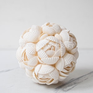 May include: A decorative sphere composed of numerous small, white seashells, arranged to resemble a flower. The sphere is set against a white background, highlighting the intricate texture and natural color of the shells. The product is from Tonkin Handicraft.