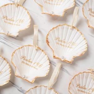 May include: White seashell ornaments with gold trim and a pearl accent. Each shell has the text "Psalm 136 His Mercy is More" printed in gold. A cream-colored ribbon is attached for hanging.