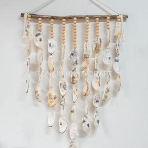 May include: A wall hanging featuring natural oyster shells suspended from wooden beads and a driftwood branch. The shells are off-white with gray and brown markings. The text "TONKIN HANDICRAFT" is visible.