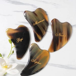 May include: Four brown and black horn facial massage tools with gold lettering. The tools are shaped like a heart and have the names Gigi, Maman, Noa, and Sophie inscribed on them.