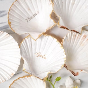 Op de afbeelding: Vijf witte schelpen met een gouden rand, elk met een naam erop geschreven. De namen zijn Vanessa, Lydia en drie anderen. De schelpen zijn gerangschikt op een wit oppervlak met een witte orchidee.