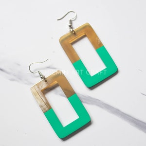 Peut inclure: Une paire de boucles d'oreilles rectangulaires avec un schéma de couleurs or et vert. Les boucles d'oreilles sont en corne et ont un design simple et moderne.