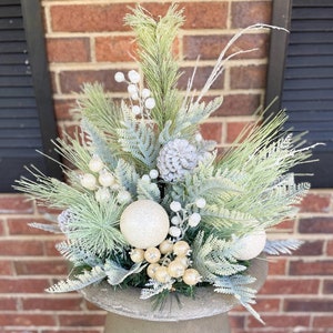 May include: A decorative outdoor urn filled with winter greenery. The arrangement includes frosted pine branches, white berries, and glittery ornaments. The urn is made of concrete and has a classic, elegant shape, perfect for a front porch or garden.