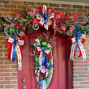 May include: A festive Christmas door decoration featuring a red door adorned with a matching garland and wreath. The garland and wreath are decorated with red, green, and blue accents, including bows and ornaments. The door is framed by a brick wall.