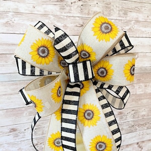 Puede incluir: Un gran lazo hecho con cinta de arpillera a rayas blancas y negras y una cinta de arpillera beige con estampado de girasol. El lazo est&aacute; atado alrededor de una linterna blanca.