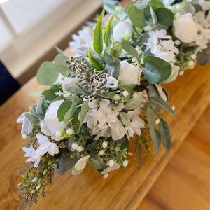 May include: A white and green floral centerpiece with white flowers and eucalyptus leaves. The centerpiece is on a wooden table.