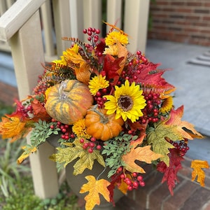 May include: A decorative urn overflowing with autumn foliage, including sunflowers, pumpkins, and colorful leaves. The arrangement features vibrant red berries and a variety of textures, creating a festive fall display. The urn is a muted green color.