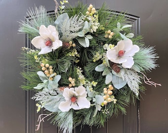 Può includere: Una ghirlanda di magnolia bianca con rami di pino verdi, bacche bianche e pigne marroni. La ghirlanda è decorata con verde ghiacciato e fiori di magnolia bianchi.