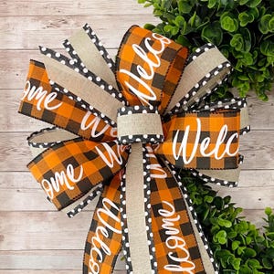 May include: A black and orange buffalo plaid ribbon with the word "Welcome" written in white on it. The ribbon is tied into a bow with a beige ribbon with white polka dots.