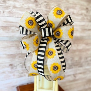 Puede incluir: Un gran lazo hecho con cinta de rayas blancas y negras y cinta de arpillera con estampado de girasol. El lazo est&aacute; atado en un gran nudo completo.