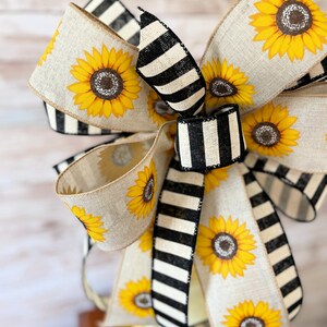 Puede incluir: Un gran lazo hecho con dos tipos de cinta. Una cinta es de arpillera natural con estampado de girasol. La otra cinta es de rayas blancas y negras. El lazo est&aacute; atado en un gran nudo completo.