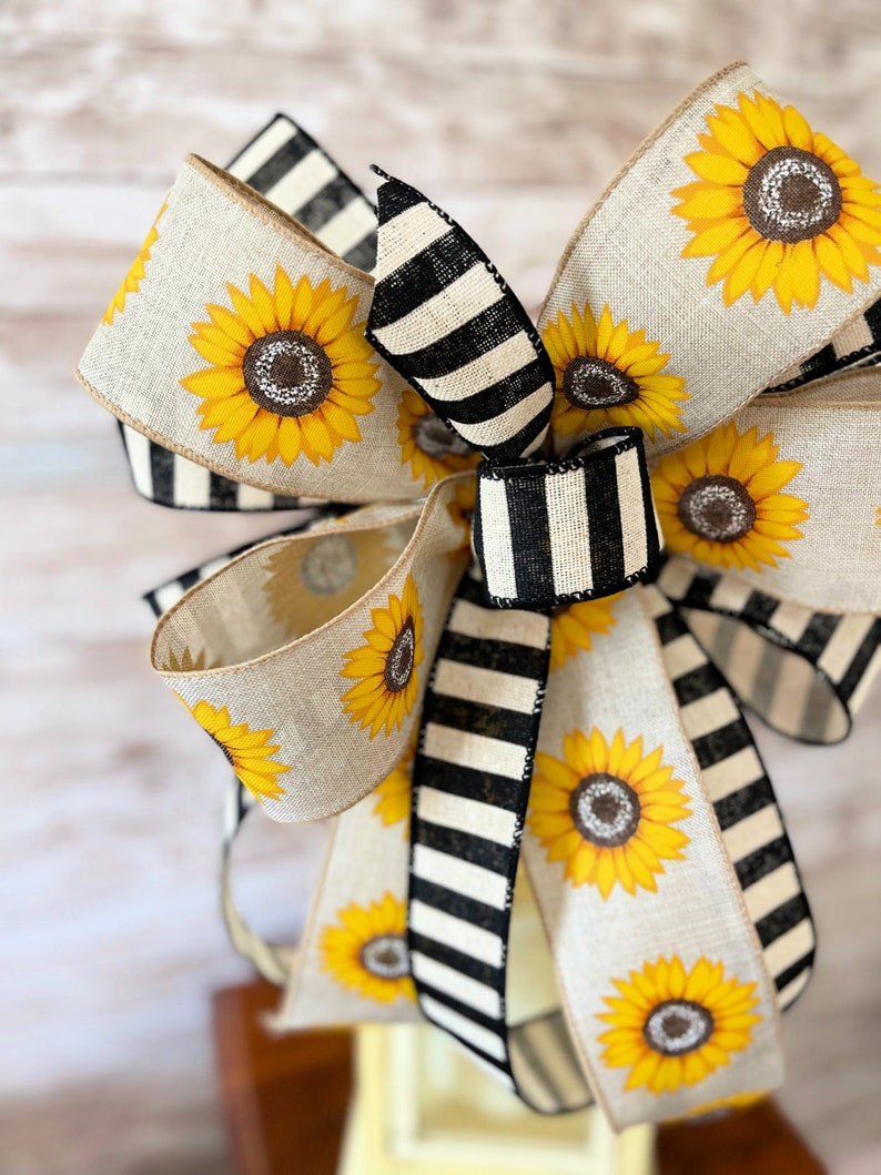 Puede incluir: Un gran lazo hecho con dos tipos de cinta. Una cinta es de arpillera natural con estampado de girasol. La otra cinta es de rayas blancas y negras. El lazo est&aacute; atado en un gran nudo completo.