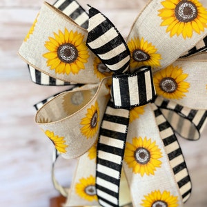 Puede incluir: Un gran lazo hecho con dos tipos de cinta. Una cinta es de arpillera natural con estampado de girasol. La otra cinta es de rayas blancas y negras. El lazo est&aacute; atado en un gran nudo completo.