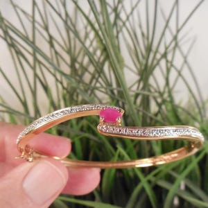 May include: A gold-toned hinged bangle bracelet featuring a bypass design with a central round pink gemstone. The two curved arms of the bracelet are accented with a row of small, clear pavé-set stones that extend along the top surface.