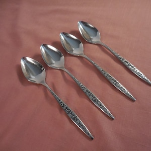 May include: Four silver spoons with textured handles are arranged diagonally on a pink surface. The spoons have a polished, reflective bowl and a detailed geometric pattern on the handle. The spoons are likely for dining.