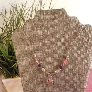 May include: A silver necklace with a heart pendant. The necklace features a strand of colorful beads in shades of pink, purple, and yellow. The heart pendant is a light pink color with a bow at the top.