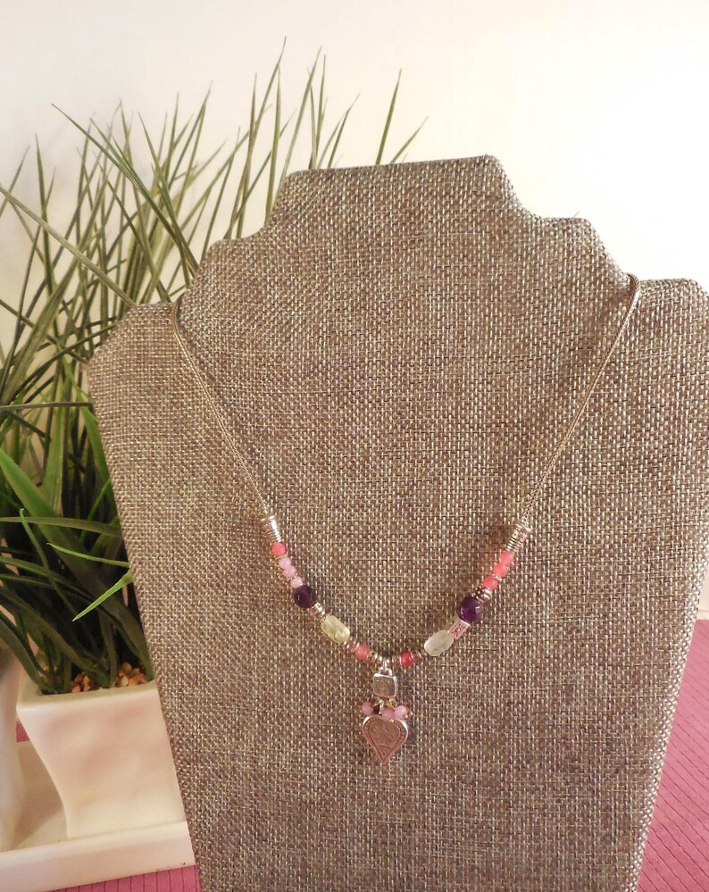 May include: A silver necklace with a heart pendant. The necklace features a strand of colorful beads in shades of pink, purple, and yellow. The heart pendant is a light pink color with a bow at the top.