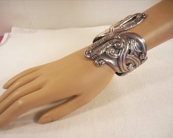 Puede incluir: Un brazalete plateado con un diseño ornamental y arremolinado. El brazalete se muestra en una mano de maniquí, destacando sus intrincados detalles y artesanía. El brazalete presenta un estilo artístico único.