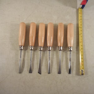 May include: Set of seven wood carving tools with light brown wooden handles and silver blades. A yellow measuring tape is positioned next to the tools. The tools are arranged in a row, showcasing different blade shapes for detailed woodworking.
