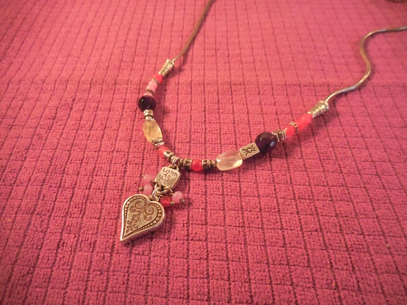 May include: A necklace with a silver heart pendant and a beaded chain. The heart pendant has an ornate design and the words "Listen to your heart". The beads are various colors, including pink, red, and clear.