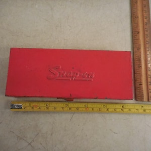 May include: A red Snap-on tool box with the brand name embossed on the lid. A wooden ruler and a yellow measuring tape are next to the box. The box is approximately 7 inches long.