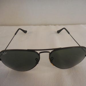 May include: Black aviator sunglasses with dark green lenses. The sunglasses have a thin, dark metal frame and a double bridge. The Ray-Ban logo is visible on the left lens. The sunglasses are a classic style.